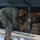 Boots in the Snow A military service member in MARPAT (Marine pattern) camouflage uniform is loading or retrieving tactical gear from the undercarriage luggage compartment of a bus or coach vehicle. Multiple tan/coyote brown military rucksacks and tactical bags are packed into the storage compartment. Snow is visible on the ground, indicating a cold-weather environment.