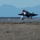 Lightning II strikes Iwakuni, F-35B arrives A fifth-generation stealth fighter jet, consistent with an F-35 Lightning II, is captured during a takeoff or landing roll on a military airstrip at dusk or dawn. The aircraft's landing light is brightly illuminated, and its landing gear is deployed. A mountainous landscape is visible in the hazy blue background.