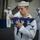 170117-N-BL637-084 A U.S. Navy sailor in dress white uniform carefully folds an American flag during what appears to be a formal military ceremony. The foreground sailor displays a Petty Officer rating badge on his left sleeve and handles the tri-folded flag with visible concentration and solemnity. Additional sailors in dress whites are visible in the background, some appearing to salute.