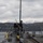 170123-N-JI086-205 A naval personnel member stands on the deck of a military vessel, positioned near what appears to be a deck-mounted weapon system. The ship is operating in calm waters with a coastal landscape visible in the background, featuring snow-capped mountains and hillside settlements. The overcast sky and winter conditions suggest cold weather operations.