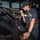 170121-N-KB426-044 A female U.S. Navy sailor is standing at a navigation/helm console on the bridge of a naval vessel, speaking into a telephone handset. She is wearing a navy blue uniform with a name tag reading 'BIG BAD JO...' and a blue cover (cap) with an anchor insignia. A second crew member is visible in the background at another station.