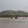 Camp Pendleton Flooding 2017 The image shows a flooded lowland area with murky brown water submerging shrubs and vegetation in the foreground. A large green hill or low mountain dominates the background, covered in lush grass likely due to recent rainfall. The overcast, gray sky and saturated landscape suggest active or recent heavy precipitation causing the flooding.