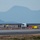 Lightning II strikes Iwakuni, F-35B arrives A military jet aircraft, appearing to be an F-35, is captured during takeoff or landing on a runway at dusk or dawn. Several personnel and a white van are visible near taxiway signage on the airfield. A mountainous landscape is visible in the hazy background.