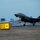 Lightning II strikes Iwakuni, F-35B arrives A dark-colored fifth-generation stealth fighter jet, consistent with an F-35 variant, is captured during takeoff or landing on a runway at dusk or dawn. The aircraft's landing gear is deployed and a landing light is illuminated on the nose gear. A yellow runway marker labeled 'B2' is visible in the foreground, with an air traffic control tower and airport structures in the background.