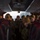 Boots in the Snow A group of military personnel in camouflage uniforms and tan/olive beanie hats are seated inside a coach bus with red seats and red curtains. The image is taken from the rear of the bus looking toward the front, with one individual in the center aisle appearing to address or observe the group. The lighting is dim with natural light coming through the front windshield.
