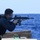 170119-N-RM689-452 A uniformed military or law enforcement personnel is firing a rifle from the deck of a naval vessel over open ocean. The individual is equipped with tactical gear and appears to be conducting a live-fire training exercise or operational drill at sea. A spent casing appears to be ejecting from the weapon, indicating active firing.
