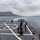 170123-N-KP948-213 The image shows the aft deck of a military naval vessel underway in coastal waters, with mountainous terrain visible in the background. Multiple crew members dressed in dark navy uniforms are gathered on the helicopter landing pad area of the stern deck. The ship appears to be departing or maneuvering, as evidenced by the visible wake in the water behind it.