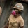 NMCB 5 Classroom Construction Project in Panay A military service member is shown wearing U.S. Navy AOR2 digital camouflage trousers and a heavily soiled tan t-shirt, indicating active labor or disaster relief work. The individual is equipped with personal protective equipment including a hard hat bearing a green safety cross emblem, a pink half-face respirator, safety glasses, and foam earplugs. The overall appearance suggests participation in construction, demolition, or humanitarian assistance operations.