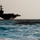 USS Carl Vinson (CVN 70) A large naval aircraft carrier is photographed in silhouette against a pale, hazy sky while underway on open, choppy blue ocean waters. The vessel is viewed from a low angle at a distance, emphasizing the scale of the ship against the rough sea. The image has a dramatic, moody quality due to the strong backlighting and dark silhouette of the ship.
