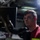 Metal Work A young male technician or mechanic wearing safety glasses and a red polo shirt is working in what appears to be a garage or maintenance bay. He is looking upward, likely inspecting the underside of a vehicle raised on a lift. The image is dramatically lit with a bright work light in the background, creating strong contrast.