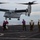 Marines return to USS Mesa Verde A V-22 Osprey tiltrotor aircraft is hovering over the flight deck of a naval vessel, appearing to be in the process of landing. Multiple deck crew members in color-coded vests and helmets are observing and managing the landing operation. The scene takes place at sea under overcast skies.
