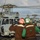 170123-N-CG677-175 Two naval deck crew members stand on a flight deck directing helicopter operations, with multiple military helicopters visible in the background. The personnel are wearing color-coded helmets and vests indicating their roles, with one helmet marked '7H' in red and the other marked 'LSE 7H' in green. The scene appears to be aboard a U.S. Navy vessel, likely an amphibious assault ship or aircraft carrier.
