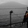 170124-N-JI086-041 A uniformed military or naval personnel stands on the deck of a vessel, holding a rifle and wearing an orange high-visibility safety vest over camouflage fatigues. The scene is set in overcast, rainy conditions on a body of water with a mountainous landmass visible in the background. A floating barrier or net line is visible stretching across the water in the mid-ground.
