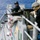USS Lake Champlain (CG 57) Crew-Served Weapons Shoot Two military personnel are positioned on the deck of a naval vessel at sea, operating a large crew-served machine gun. One sailor wears a black cap with headset and sunglasses, while the other wears a ballistic helmet, face covering, and body armor marked 'NAVY'. The ship's hull displays colorful signal flag decorations, and open ocean is visible in the background.