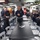 170117-N-QF605-105 A group of naval personnel in dark blue coverall uniforms are gathered in what appears to be a mess hall or briefing room aboard a military vessel. A senior officer or NCO is standing and addressing the assembled sailors, with two individuals facing him in a briefing or discussion posture. The setting features institutional furnishings typical of a naval ship interior.
