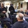 170117-N-QF605-075 A group of U.S. Navy personnel in dark blue coveralls are gathered in what appears to be a Chief Petty Officers' mess aboard a naval vessel. One individual is standing and addressing the group, holding a white coffee mug, while others are seated around a table. The room is decorated with Navy memorabilia, plaques, a framed American flag, and a 'Yuengling' branded poster.