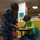 NPASE COMREL A U.S. Navy service member in blue digital camouflage (NWU Type I) uniform, with the name tape 'PATTEN' visible, is leaning over to assist a young boy seated at a school desk. The boy, wearing a green t-shirt, is engaged in a coloring or drawing activity on a sheet of paper. The setting appears to be a classroom or educational facility, likely on a military installation.