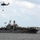 Vertical Replenishment A large US Navy amphibious assault ship, bearing the hull number '5' on its island superstructure, is anchored or underway at sea. Two military helicopters are visible — one performing a vertical replenishment (VERTREP) operation with a sling load suspended beneath it, and a second helicopter flying near the ship's port side. The flight deck is populated with multiple rotary-wing aircraft and equipment.