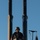 USS Cheyenne hosts bussines leaders The image shows U.S. Navy personnel stationed on what appears to be the sail (conning tower) of a submarine. One sailor in the foreground wears a tactical helmet and body armor with visible text reading 'US NAVY,' and appears to be armed. An American flag is visible on the right side of the frame, and two large mast/antenna structures rise into a clear blue sky.