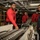 170118-N-BL637-022 A group of individuals in a military/naval setting, several wearing red sweatshirts with black markings, are gathered around what appears to be an ammunition belt or weapons system on a table. The setting appears to be the interior of a naval vessel, identifiable by the overhead piping, fluorescent lighting, and industrial structure. One person in a red sweatshirt labeled 'G-3' appears to be explaining or demonstrating the equipment to the group.