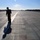 Every clime and place: VMAT-203 Marines maintain expeditionary mindset at MCALF Bogue A person in a dark flight suit and helmet stands on a large concrete runway or taxiway at an airfield, backlit by the sun creating a strong silhouette effect. The wide expanse of concrete extends toward the horizon, flanked by tree lines on both sides. The low sun angle casts a long shadow of the figure across the runway surface.