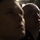 USS Cheyenne hosts bussines leaders A dramatic, low-light close-up photograph of two individuals in the foreground, with at least one wearing what appears to be a military or naval uniform with insignia visible on the collar. The person in the middle ground is bald, wearing rectangular-framed glasses, and appears to be looking upward. The image uses strong chiaroscuro lighting, creating deep shadows across the subjects' faces.