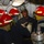 170117-N-OA516-0026 A uniformed naval officer wearing a cap with 'Newport, Florida' text is smiling and interacting with three personnel wearing red firefighting/damage control helmets and protective gear in what appears to be a ship's interior compartment. The setting shows a confined mechanical space with pipes, cables, and equipment typical of a naval vessel. The scene appears to depict a damage control training or inspection scenario.