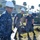 170120-N-AW702-003 A group of U.S. Navy personnel wearing Navy Working Uniform (NWU) digital camouflage and white hard hats are engaged in a logistics or setup operation outdoors on what appears to be a military installation. The foreground shows three sailors carrying metal equipment, likely folding tables or frames, while numerous other personnel work in the background. The setting includes palm trees and institutional buildings consistent with a coastal military base.