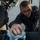 170123-N-WE240-040 A young military servicemember in a dark navy uniform is performing maintenance work on what appears to be a military vehicle or weapons system. He is leaning into a confined mechanical space, wiping down components with a blue cloth. His uniform displays rank insignia and patches consistent with U.S. Navy enlisted personnel.