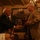 Ken Pipes Retirement Ceremony Two older men are shaking hands in what appears to be a restaurant or private dining room with warm, dim lighting. One man wears a dark navy blazer and the other wears a tan/beige suit jacket, and the man in the tan jacket appears to be holding a red box or gift. The setting includes framed artwork, an American flag visible in the background, and decorative string lights.