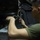 From bolts to bombs A military service member in an olive green t-shirt is working with rope, tying or securing a knot inside what appears to be a military vehicle or aircraft interior. The individual is focused on manipulating a length of natural-fiber rope connected to a carabiner or clip above. A metal box or container marked '0342 TAM' is visible in the foreground.