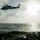 USS Lake Champlain (CG 57) Flight Quarters A military helicopter with a distinctive camouflage paint scheme hovers above the flight deck of a naval warship at sea. The image is taken from the perspective of someone aboard the ship, looking out toward the open ocean with the sun partially obscured by clouds. The scene depicts an active naval aviation operation, likely a landing or hover exercise.