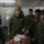 2d Medical Bn Field Exercise Military medical personnel are conducting what appears to be a combat casualty care training exercise inside a field medical tent. Several individuals in military camouflage uniforms, including one wearing a stethoscope, are gathered around a simulated patient on a stretcher with mock wounds and medical supplies. A civilian instructor wearing a cap labeled 'INSTRUCTOR' observes and evaluates the exercise while holding a clipboard.