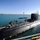 Expeditionary ordnance loading exercise A large military submarine is docked at a naval facility, with personnel on its deck overseeing a crane-assisted loading or unloading operation of what appears to be a torpedo or similar cylindrical weapon/equipment. The submarine is dark-colored with a visible sail (conning tower) and is moored alongside a pier in calm, blue-green waters. Approximately 15-20 personnel in various uniforms are visible on the hull.