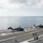 170118-N-NV908-045 The image shows the flight deck of a US Navy aircraft carrier with helicopter operations underway. Two MH-60 Seahawk helicopters are on the deck with rotors spinning, while a third helicopter is airborne in the background. Multiple flight deck crew members in color-coded jerseys are visible managing operations.