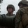Artillery Training School Two military personnel in combat gear are positioned near a large weapon system, likely a recoilless rifle or anti-tank weapon. Both individuals are wearing MARPAT (Marine pattern) camouflage uniforms and combat helmets, suggesting they are U.S. Marines. The image appears to capture a training or operational scenario with the weapon barrel prominently visible in the foreground.