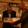 Ken Pipes Retirement Ceremony A U.S. Marine Corps officer in dress uniform is presenting a Certificate of Commendation to a civilian man. The ceremony appears to be taking place in a room decorated with military memorabilia, including a Map of Korea on the wall behind them. Both individuals are looking down at the open presentation folder containing the certificate.