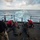 170123-N-VN584-328 Military personnel are conducting a live-fire weapons exercise on the stern deck of a naval vessel at sea. Several sailors in prone and kneeling positions are operating crew-served weapons, with supervisors in red shirts and a safety observer wearing a cap marked 'WTT' overseeing the drill. Spent brass casings are visible on the deck, and the ship's wake is clearly visible in the open ocean behind them.