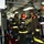 161229-N-N0901-778 A group of firefighters or damage control personnel are assembled in a narrow corridor, likely aboard a naval vessel. They are equipped with INTERSPIRO self-contained breathing apparatus (SCBA) tanks, red helmets, and fire-resistant suits with high-visibility yellow reflective strips. The setting and equipment suggest a firefighting drill or emergency response exercise.