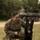 31st Marine Expeditionary Unit completes MEUEX A U.S. Marine in MARPAT camouflage uniform, identified by the name tape 'MARIANI,' kneels beside a black German Shepherd military working dog on a grassy field. The Marine is smiling and holding the dog's leash while the dog sits attentively with its tongue out. In the background, a military wheeled armored vehicle (likely an LAV or Stryker) is visible with personnel on top.