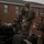Artillery Training School Two military personnel in camouflage uniforms and helmets are working on what appears to be a towed artillery piece, likely an M777 howitzer or similar field gun. The activity takes place in front of a two-story brick building, suggesting a military base or installation. One soldier is focused on the gun's sighting mechanism while the other appears to be handling a component in motion.