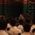Japan Ground Self-Defense Force tours Marine Logistics Group facilities A military personnel in camouflage uniform appears to be conducting a briefing or presentation to a large group of individuals dressed in white shirts inside what appears to be a warehouse or storage facility. The audience members are seated with their backs to the camera, facing the speaker. The setting suggests a formal instruction or orientation session in an industrial or logistics environment.