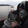 170123-N-KP948-067 A naval or maritime personnel member is using a gyrocompass or bearing repeater instrument on the bridge wing of a vessel. The individual is wearing military-style communication headphones and a dark beanie cap with partial text visible reading 'DI G...' The scene is set in a coastal waterway with at least one tugboat visible in the background.