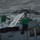 170123-N-VN584-172 Two naval aviation technicians in green jerseys perform maintenance on a military helicopter, likely an MH-60 Seahawk, aboard a naval vessel at sea. The personnel have opened an access panel on top of the aircraft to inspect or service internal components. The rough sea conditions visible in the background suggest active underway operations.