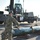 Expeditionary ordnance loading exercise Military personnel in camouflage uniforms and hard hats are handling a large green torpedo or underwater weapon on a pier or dock area. A heavy-lift crane vehicle is positioned in the background, suggesting an ordnance loading or transfer operation. The setting appears to be a naval facility during daylight hours.