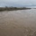Camp Pendleton Flooding 2017 A wide, fast-moving river in flood stage with heavily turbulent, sediment-laden brown water. The surrounding landscape shows bare, dormant riparian vegetation under an overcast, stormy sky. The scene indicates recent or ongoing heavy rainfall causing elevated water levels and strong currents.