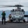 170117-N-BL637-205 A U.S. Navy helicopter, marked with the number '610', is on the flight deck of a naval vessel with rotors spinning. Several deck crew members in colored vests and helmets are positioned around the aircraft, performing pre-flight or post-flight operations. A mountainous coastline, consistent with Hawaii, is visible in the background across open water.