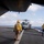 COMPTUEX VertRep The image shows a flight deck operations scene aboard a U.S. Navy amphibious ship, with a military helicopter (appears to be an MH-60 Seahawk, numbered 65) landed or preparing to depart on the deck. Two flight deck crew members in yellow jerseys and helmets are visible, monitoring the helicopter operations. The scene is set at sea under partly cloudy skies.