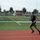 New Years Resolutions A young male athlete is running at high speed on a red synthetic track surrounding an American football field. The image uses a panning photography technique, keeping the runner relatively sharp while blurring the background to convey motion and speed. The setting appears to be a high school or community sports complex with palm trees and bleachers visible in the background.