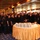 170116-N-N0901-001 A large group of people, predominantly dressed in dark/black uniforms, are gathered around a round table with a gold satin tablecloth in what appears to be a ship's dining or event hall. The table holds stacked white plates and what appears to be a cake, suggesting a celebratory event. The venue features warm amber lighting, a patterned blue carpet, and a metallic ceiling grid consistent with a cruise ship or naval vessel interior.